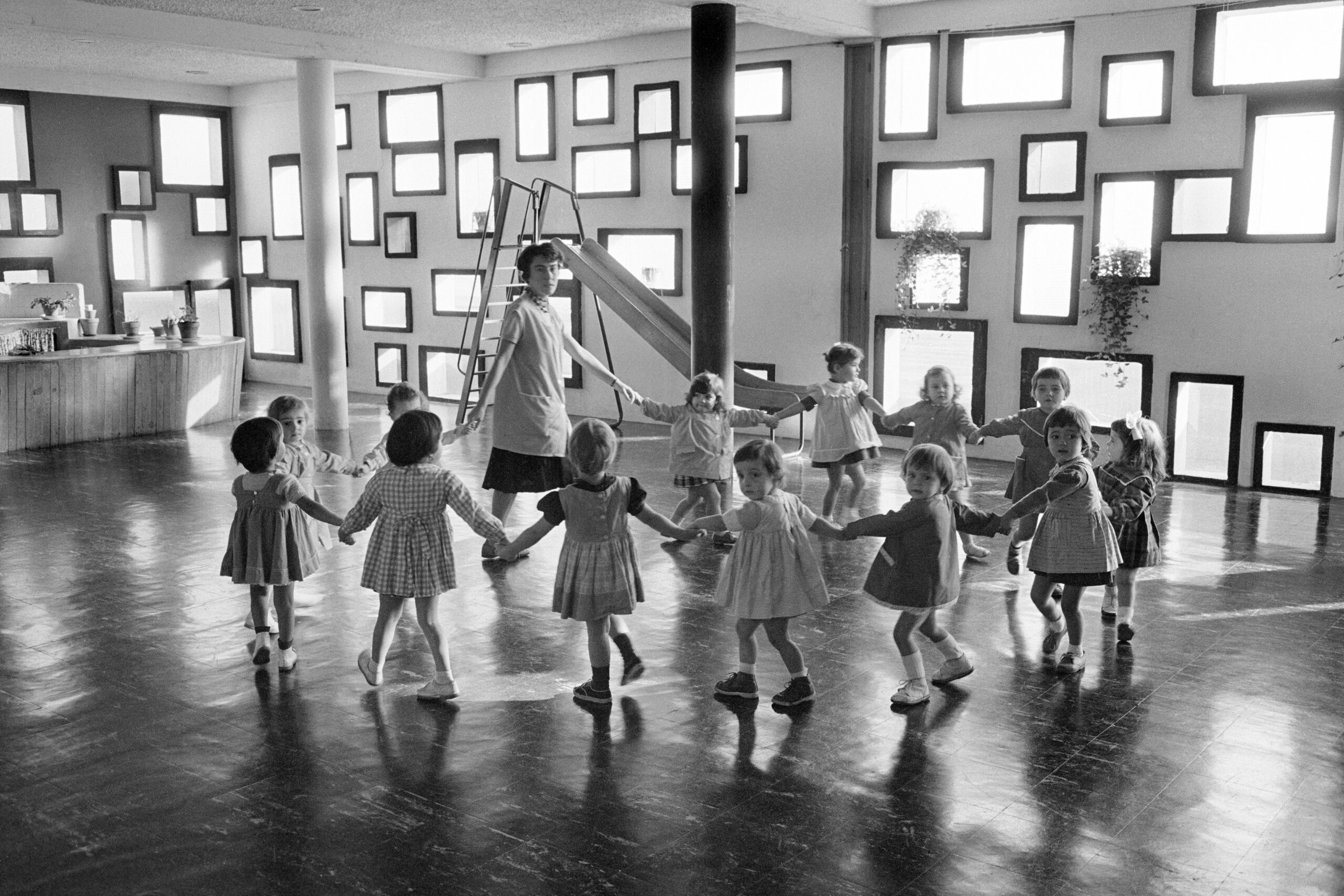 Photographie noir et blanc figurant au premier plan une ronde d'enfants et une adulte dans un intérieur avec parquet au sol et de nombreuses fenêtre sur les murs