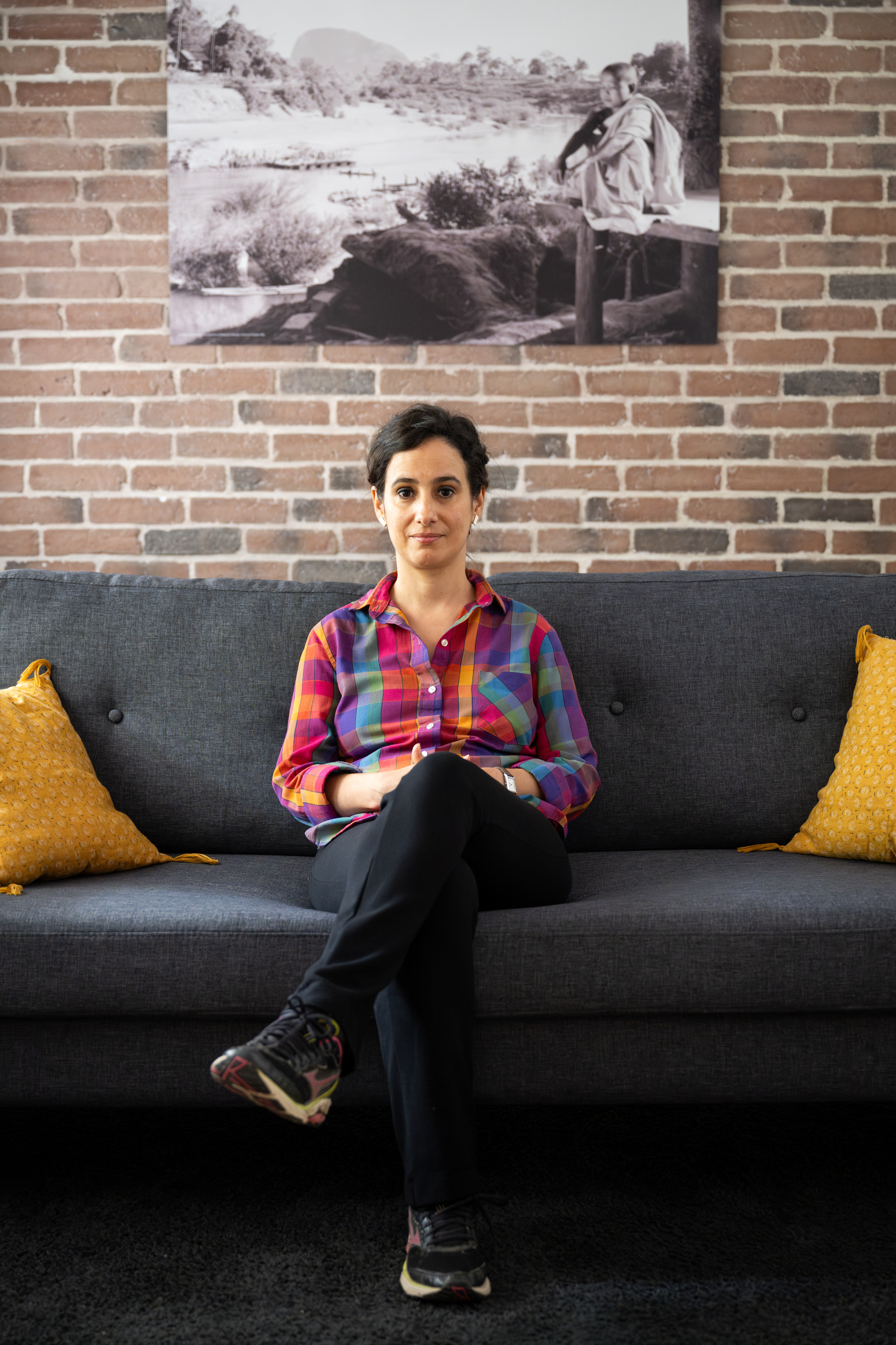 Thissa d'Avila Bensalah assise sur un canapé devant un mur de briques où est accrochée une photographie en noir et blanc